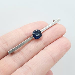 以下が含まれることがあります： 中央に4本の爪で留められた、ファセットカットが施された深い青色の正方形の石が特徴のシルバー調のバーブローチ。金属製の水平なバーは滑らかで光沢のある仕上げになっており、シンプルで直線的なデザインが施されています。