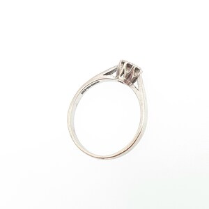 Puede incluir: Un anillo de diamantes solitario de plata. El anillo tiene un solo diamante de talla cuadrada sujeto por cuatro garras. La banda es delgada y se estrecha hacia el diamante. El anillo se muestra sobre un fondo blanco.