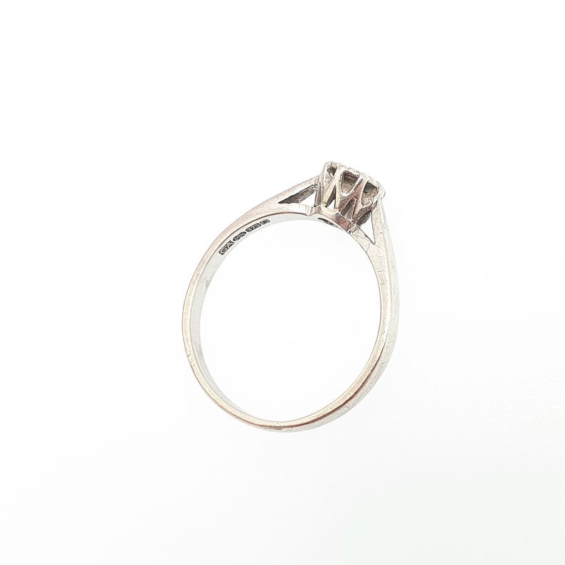 Puede incluir: Un anillo de diamantes solitario de plata. El anillo tiene un solo diamante de talla cuadrada sujeto por cuatro garras. La banda es delgada y se estrecha hacia el diamante. El anillo se muestra sobre un fondo blanco.