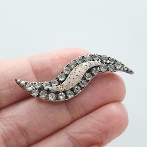 Puede incluir: Broche vintage plateado con un diseño ondulado. El broche está incrustado con numerosas piedras claras y brillantes, y presenta un acento central adornado en tono dorado. Una joya clásica.