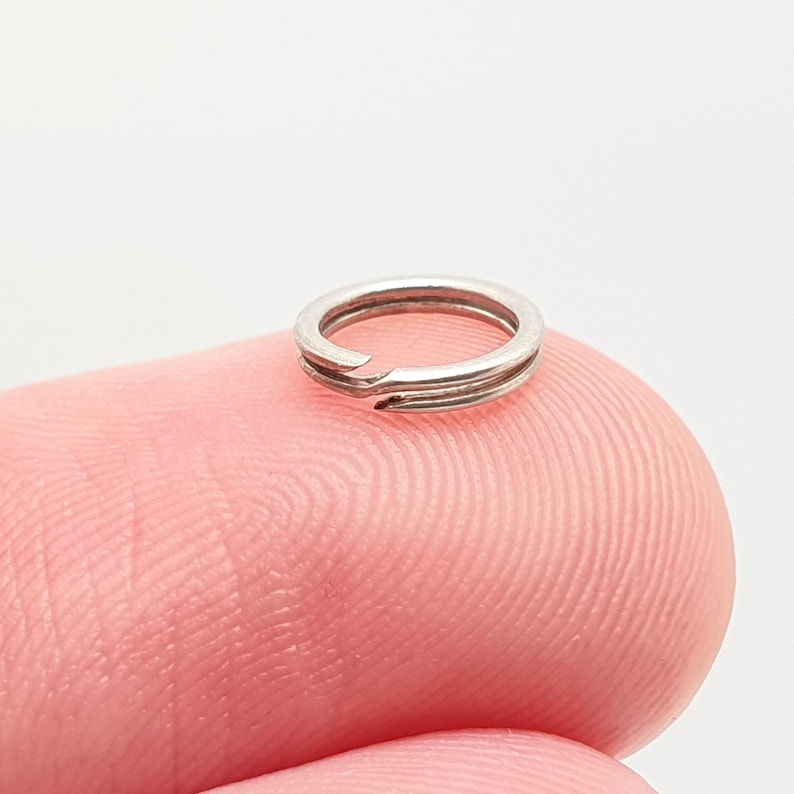 May include: A small, silver split ring with a double-loop design. The ring is open at one point, allowing for the attachment of other items. The split ring is shown on a pink fingertip.