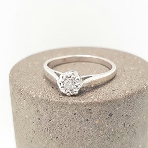 Puede incluir: Un anillo plateado con una gema clara y redonda en el centro, rodeada por un dise&ntilde;o floral. El anillo est&aacute; colocado sobre una superficie cil&iacute;ndrica gris, sobre un fondo blanco. La banda del anillo es delgada y sencilla.