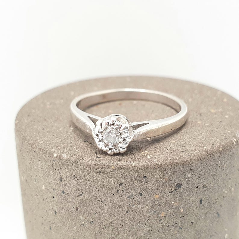 Puede incluir: Un anillo plateado con una gema clara y redonda en el centro, rodeada por un dise&ntilde;o floral. El anillo est&aacute; colocado sobre una superficie cil&iacute;ndrica gris, sobre un fondo blanco. La banda del anillo es delgada y sencilla.