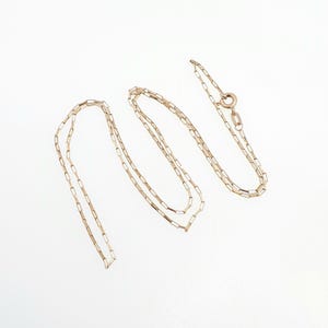 May include: A delicate gold chain necklace featuring elongated rectangular links. The chain is arranged in a loose, folded configuration and includes a circular spring ring clasp mechanism at one end, showcasing a polished metallic finish throughout.