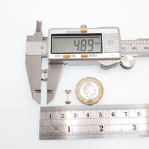 May include: A pair of small round stud earrings with a metallic finish. One earring is positioned between the jaws of a digital caliper displaying a measurement of 4.89 mm. A British one pound coin and a metal ruler are placed next to the earrings.