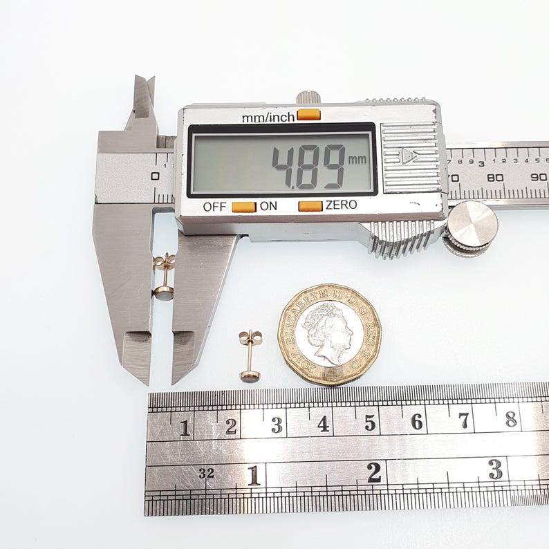 May include: A pair of small round stud earrings with a metallic finish. One earring is positioned between the jaws of a digital caliper displaying a measurement of 4.89 mm. A British one pound coin and a metal ruler are placed next to the earrings.
