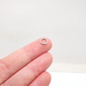 May include: A tiny, silver jump ring, held on a fingertip. The ring is circular and made of metal. The background is a plain, light colour.