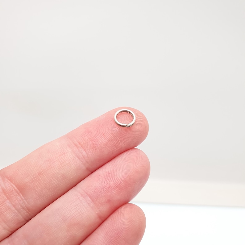 May include: A tiny, silver jump ring, held on a fingertip. The ring is circular and made of metal. The background is a plain, light colour.