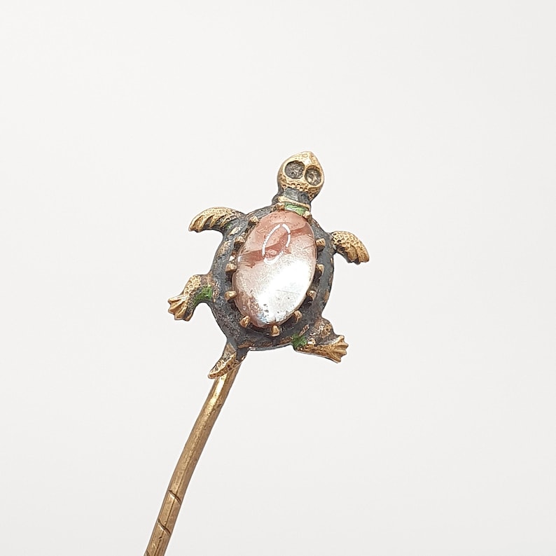 May include: A decorative hair pin featuring a turtle design. The turtle has a bronze-colored body with green accents and a central oval-shaped stone with red and white patterns. The pin's stem is a golden color.
