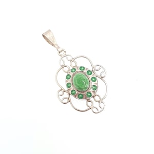 May include: A silver pendant with an oval, light green center stone surrounded by smaller green accents. The pendant has an ornate, openwork design with a loop at the top for a chain.