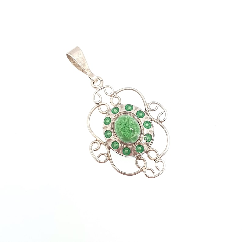 May include: A silver pendant with an oval, light green center stone surrounded by smaller green accents. The pendant has an ornate, openwork design with a loop at the top for a chain.