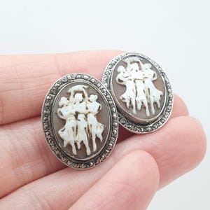 May include: A pair of oval-shaped silver earrings featuring a carved cameo design of three figures dancing in white against a brown background. The border of each earring is set with small, dark, faceted marcasite stones in a silver frame.
