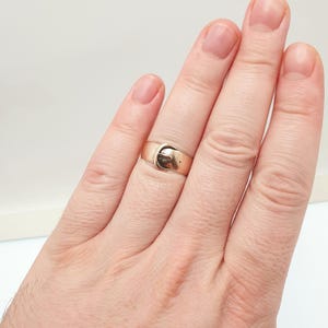 May include: A gold-colored ring with a buckle design. The ring is a band style with a small buckle detail on the front. The ring is worn on a finger, and the design is visible.