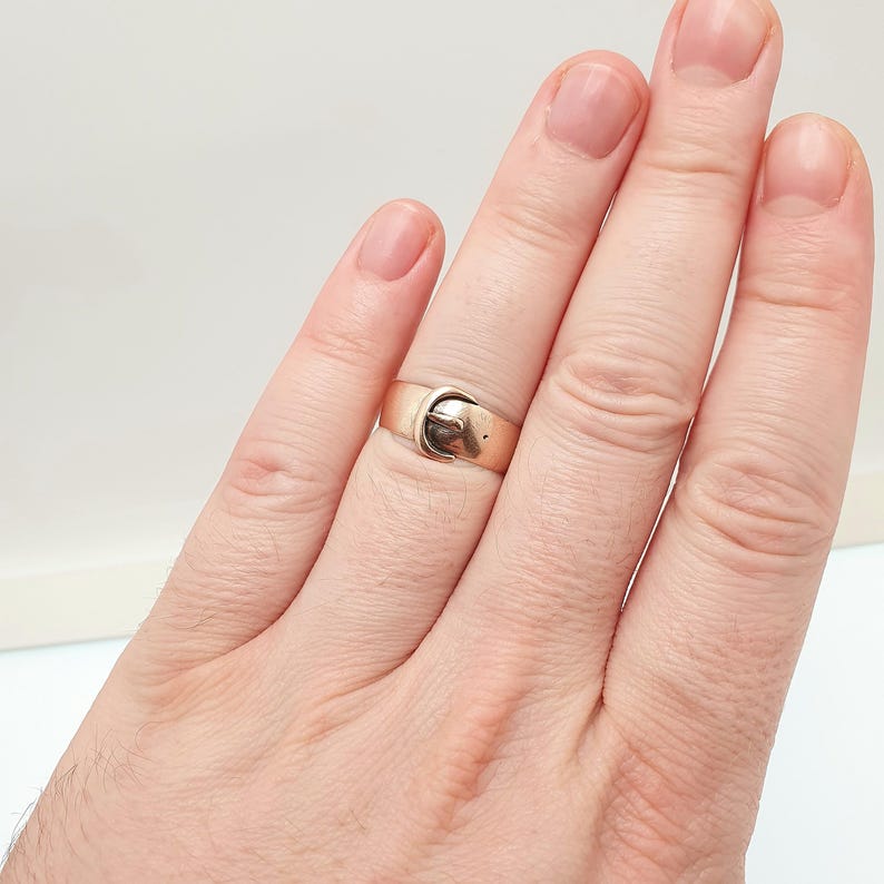 May include: A gold-colored ring with a buckle design. The ring is a band style with a small buckle detail on the front. The ring is worn on a finger, and the design is visible.