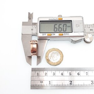 May include: A rose gold ring is measured with a digital caliper, displaying 6.60 mm. A ruler shows inches. A coin is next to the ring. The ring has a decorative top.