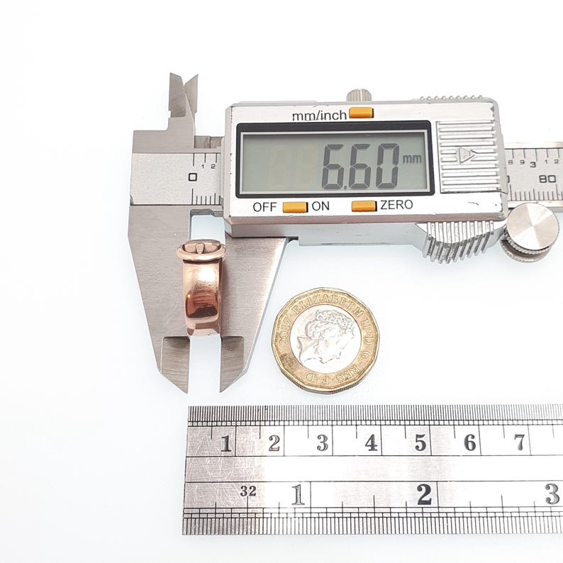 May include: A rose gold ring is measured with a digital caliper, displaying 6.60 mm. A ruler shows inches. A coin is next to the ring. The ring has a decorative top.