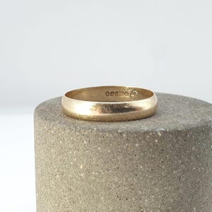 以下が含まれることがあります： 滑らかで光沢のある表面を持つゴールドのバンドリング。リングの内側には、金属の純度と真正性を示す小さな刻印やシンボルが並んでおり、リングの内周に沿って確認することができます。