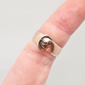 May include: A gold-colored ring with a buckle design. The ring is worn on a finger and features a detailed buckle clasp. The metal has a polished finish, reflecting light. The ring's design is a classic and timeless style.