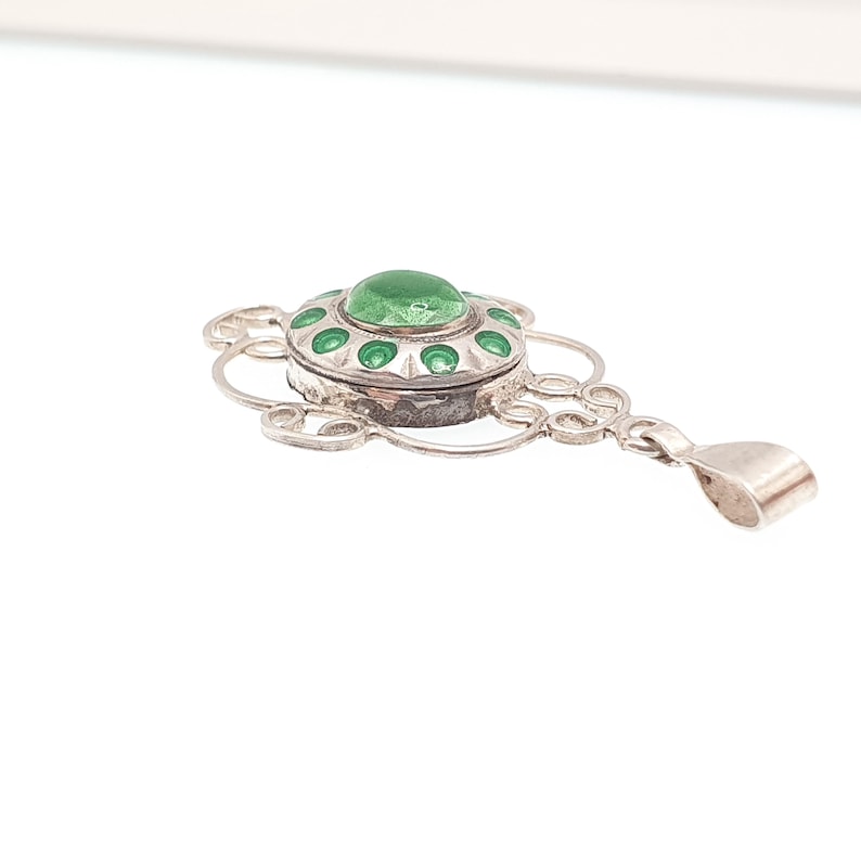 May include: A silver pendant with a central green stone and green enamel accents. The pendant has an ornate, openwork design with a bail for a chain. The piece is likely vintage or antique.