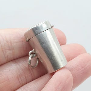 Puede incluir: Un pequeño dije cilíndrico de plata con forma de recipiente con tapa. El objeto presenta un acabado metálico liso e incluye una argolla circular unida al lateral, lo que permite colgarlo de una cadena o pulsera como un colgante.