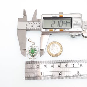 May include: A silver pendant with a green gemstone center and surrounding green accents. The pendant is measured with a digital caliper, showing a measurement of 21.04 mm. A coin is placed next to the pendant for size comparison.