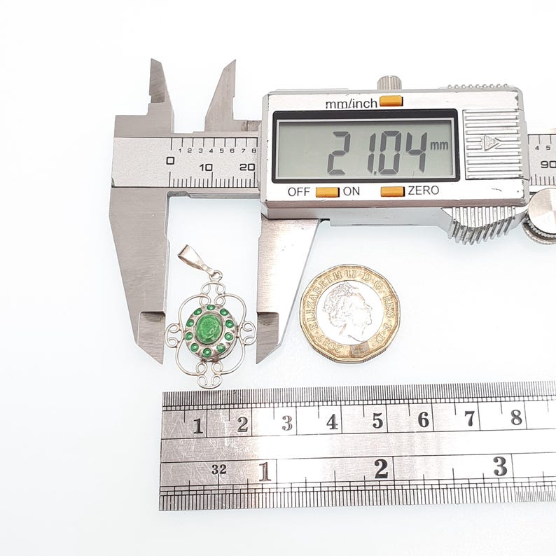 May include: A silver pendant with a green gemstone center and surrounding green accents. The pendant is measured with a digital caliper, showing a measurement of 21.04 mm. A coin is placed next to the pendant for size comparison.
