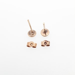 May include: A pair of rose gold stud earrings with straight posts and matching butterfly earring backs. The metal has a warm, metallic pinkish hue, and the components are displayed separately to show the post and the secure friction back mechanism.