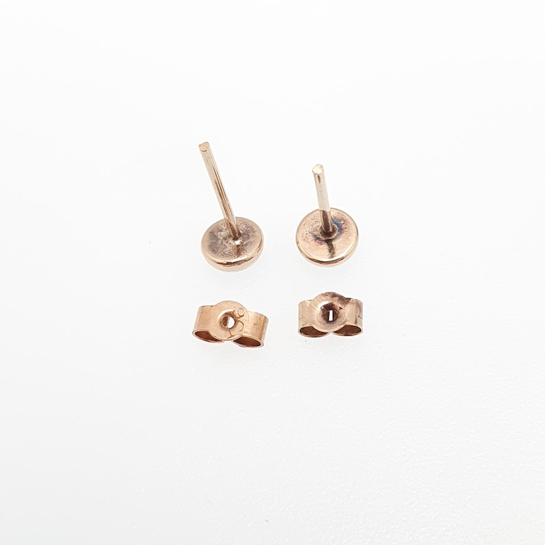 May include: A pair of rose gold stud earrings with straight posts and matching butterfly earring backs. The metal has a warm, metallic pinkish hue, and the components are displayed separately to show the post and the secure friction back mechanism.