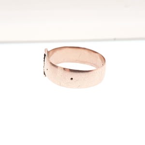 May include: A rose gold ring with a textured surface. The ring has a small decorative element and two small holes. The ring is shown on a white background.