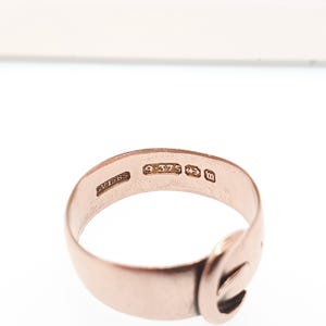 May include: A rose gold ring with a buckle design. The ring has several markings, including "9.375" and "m". The ring is set against a white background.