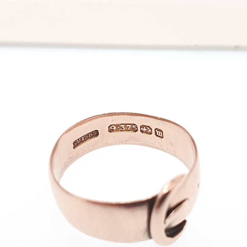 May include: A rose gold ring with a buckle design. The ring has several markings, including "9.375" and "m". The ring is set against a white background.