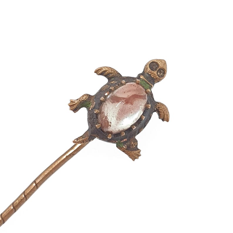 May include: A bronze-toned turtle-shaped pin with a central oval-shaped stone. The turtle has a dark brown shell with green accents and a detailed head and limbs. The pin has a long, thin, bronze-colored stem.
