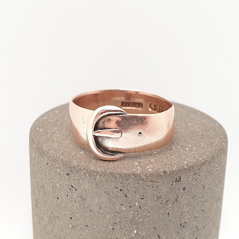 May include: A rose gold ring with a buckle design. The ring is set on a gray surface. The buckle has a curved shape with a small pin. The ring has a polished finish and a vintage aesthetic.