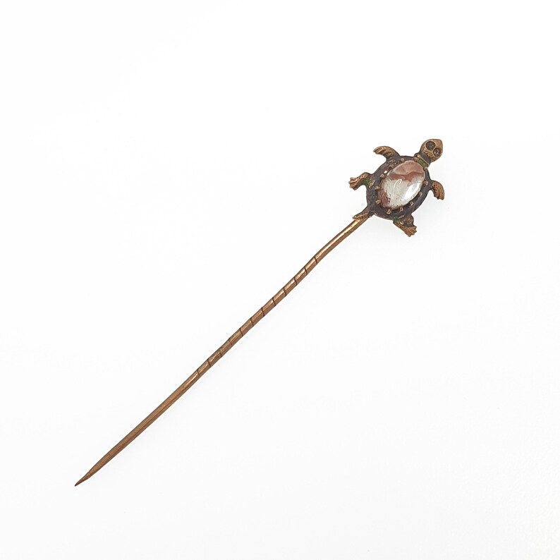 May include: A vintage hat pin with a bronze-colored metal shaft and a turtle-shaped head. The turtle has a light-colored stone set in its shell. The pin is designed to be inserted into a hat or other fabric.