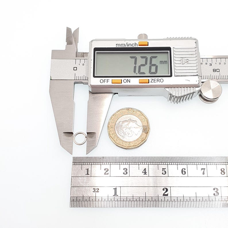 May include: A silver jump ring is measured with a digital caliper, displaying 7.26 mm. A ruler shows inches and a coin is next to the ring. The caliper reads "mm/inch", "OFF", "ON", and "ZERO".