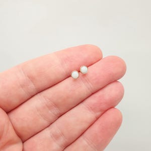 May include: A pair of small stud earrings featuring round, smooth white cabochons set in simple gold-toned metal bezels. The earrings have a minimalist circular design with a polished finish, showcasing a clean and classic aesthetic for everyday wear.