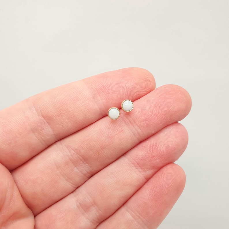 May include: A pair of small stud earrings featuring round, smooth white cabochons set in simple gold-toned metal bezels. The earrings have a minimalist circular design with a polished finish, showcasing a clean and classic aesthetic for everyday wear.
