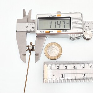 May include: A vintage turtle-shaped pin, with a pearl-like center, is measured with a digital caliper. The measurement reads 11.47 mm. A coin and ruler are also in the frame, providing scale. The pin is on a white background.