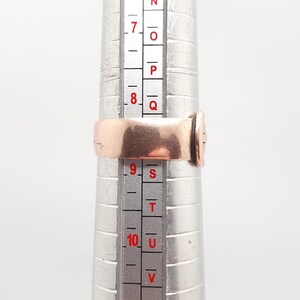 May include: A copper-colored ring is placed on a ring sizer. The ring is a simple band design. The ring sizer is silver and has measurements in inches. The ring is sized between a size 8 and 9.
