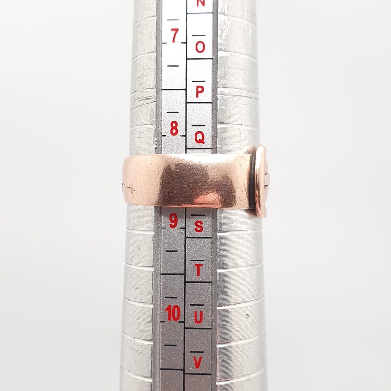 May include: A copper-colored ring is placed on a ring sizer. The ring is a simple band design. The ring sizer is silver and has measurements in inches. The ring is sized between a size 8 and 9.