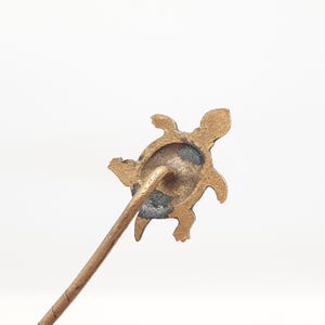 May include: A bronze-colored turtle-shaped hat pin. The pin features a detailed turtle design with a textured surface. The pin's stem is a light brown color. The background is white.