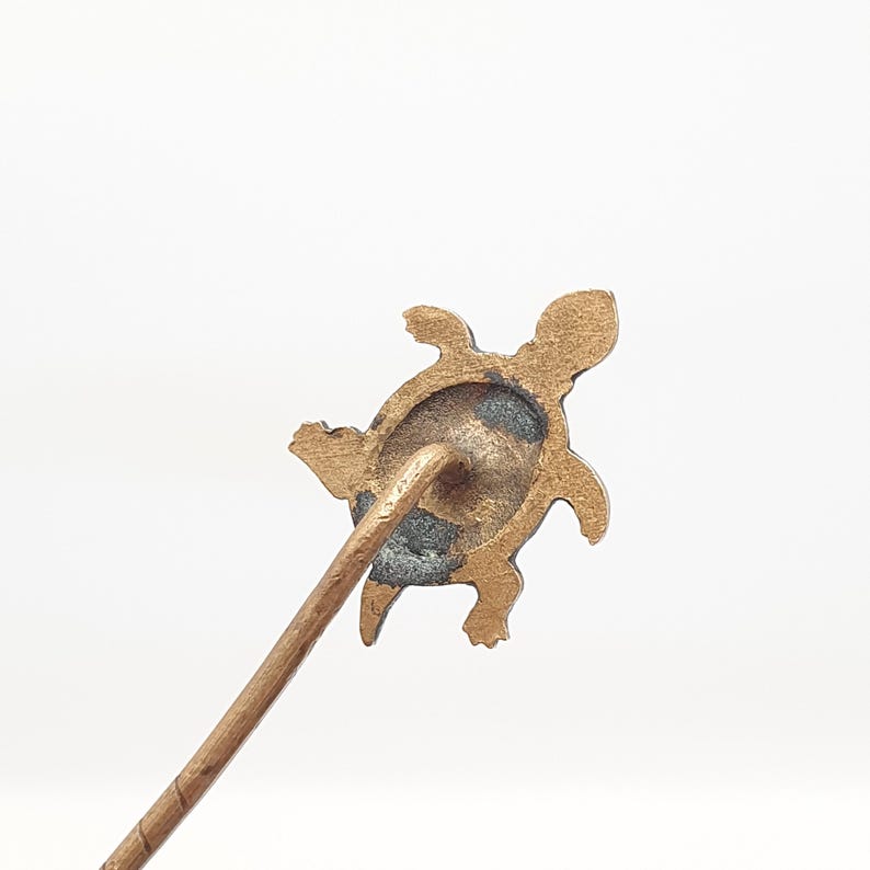 May include: A bronze-colored turtle-shaped hat pin. The pin features a detailed turtle design with a textured surface. The pin's stem is a light brown color. The background is white.