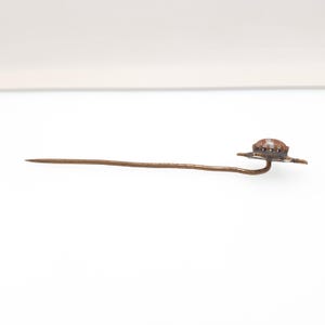May include: A vintage hat pin with a decorative hat-shaped head. The pin is made of a bronze-colored metal and features a long, slender shaft. The hat has a textured surface and is slightly domed.