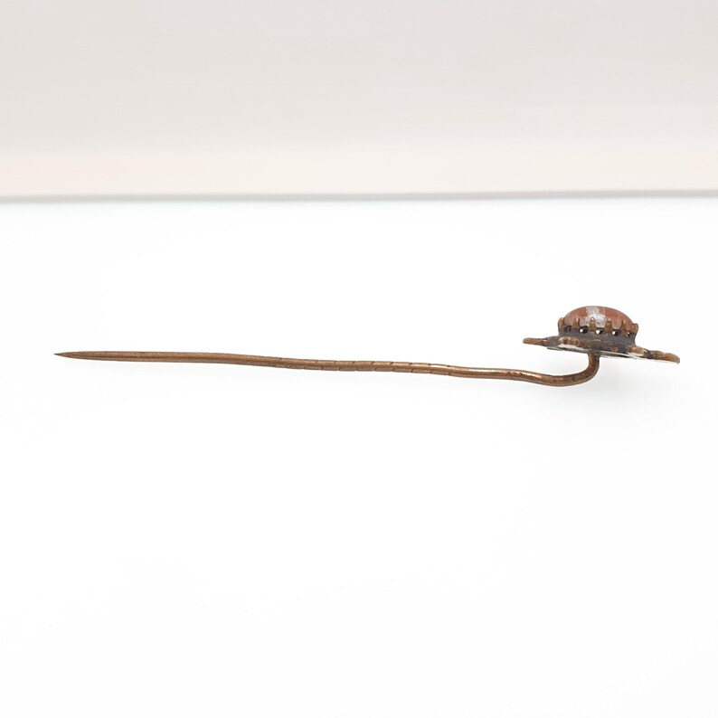 May include: A vintage hat pin with a decorative hat-shaped head. The pin is made of a bronze-colored metal and features a long, slender shaft. The hat has a textured surface and is slightly domed.