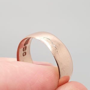 May include: A rose gold ring with a smooth, rounded band. The ring has visible markings on the inside and some surface wear. The ring is held between fingers against a neutral background.