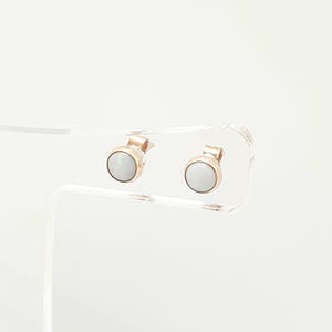 May include: A pair of stud earrings featuring round, white, iridescent opal cabochons set in polished gold-toned metal bezels. The earrings are displayed on a clear acrylic stand, showcasing the smooth, circular design and the subtle play of light across the surface of the stones.