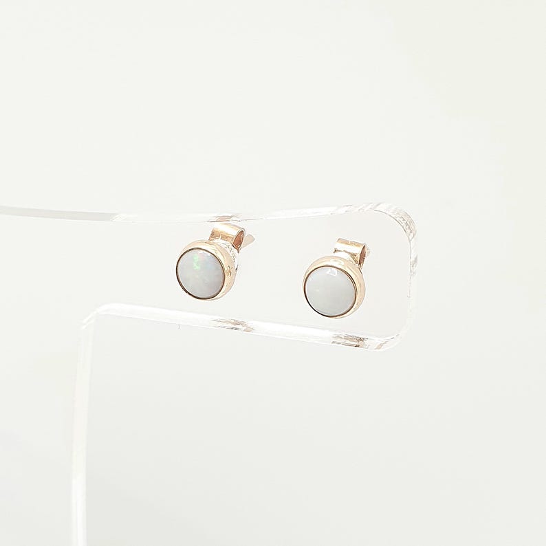 May include: A pair of stud earrings featuring round, white, iridescent opal cabochons set in polished gold-toned metal bezels. The earrings are displayed on a clear acrylic stand, showcasing the smooth, circular design and the subtle play of light across the surface of the stones.