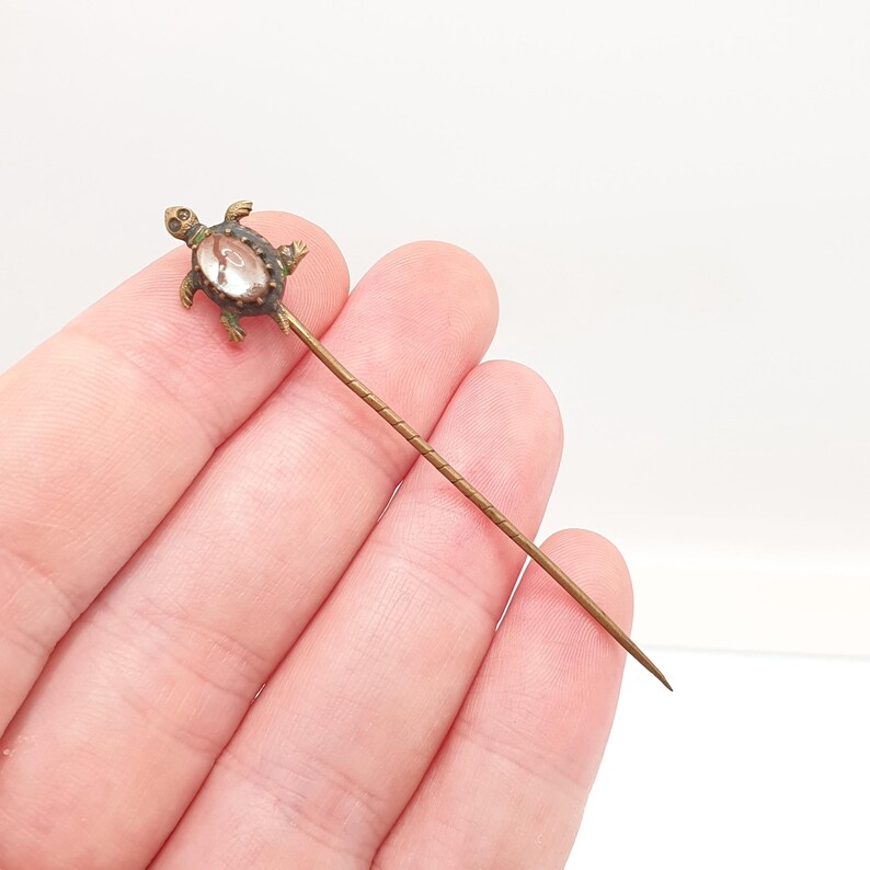 May include: A vintage hat pin with a turtle-shaped head. The turtle is made of gold-toned metal and features a clear, oval-shaped stone. The pin is long and slender, with a textured design.