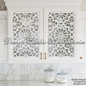 May include: White kitchen cabinets with a geometric cutout design on the doors. The cabinets have gold-coloured handles. The cabinets are above a white countertop with two glass jars on it.