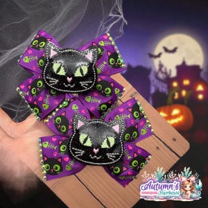 Puede incluir: Dos lazos para el pelo con temática de Halloween. Cada lazo presenta una cara de gato negro con purpurina y ojos verdes, sobre una cinta morada estampada con gatos negros y espinas de pescado. Los lazos están adornados con pedrería.